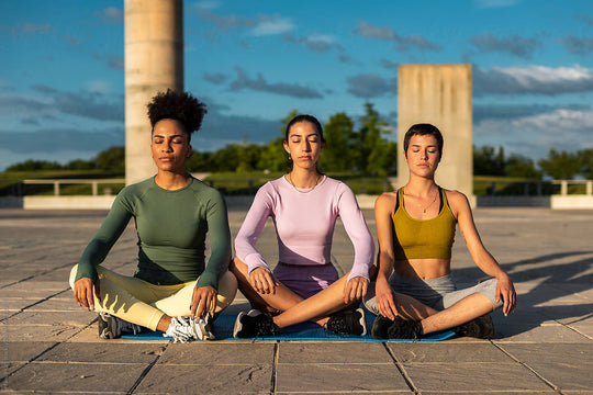 Three women sitting outside in the sun, with legs crossed and eyes closed meditation