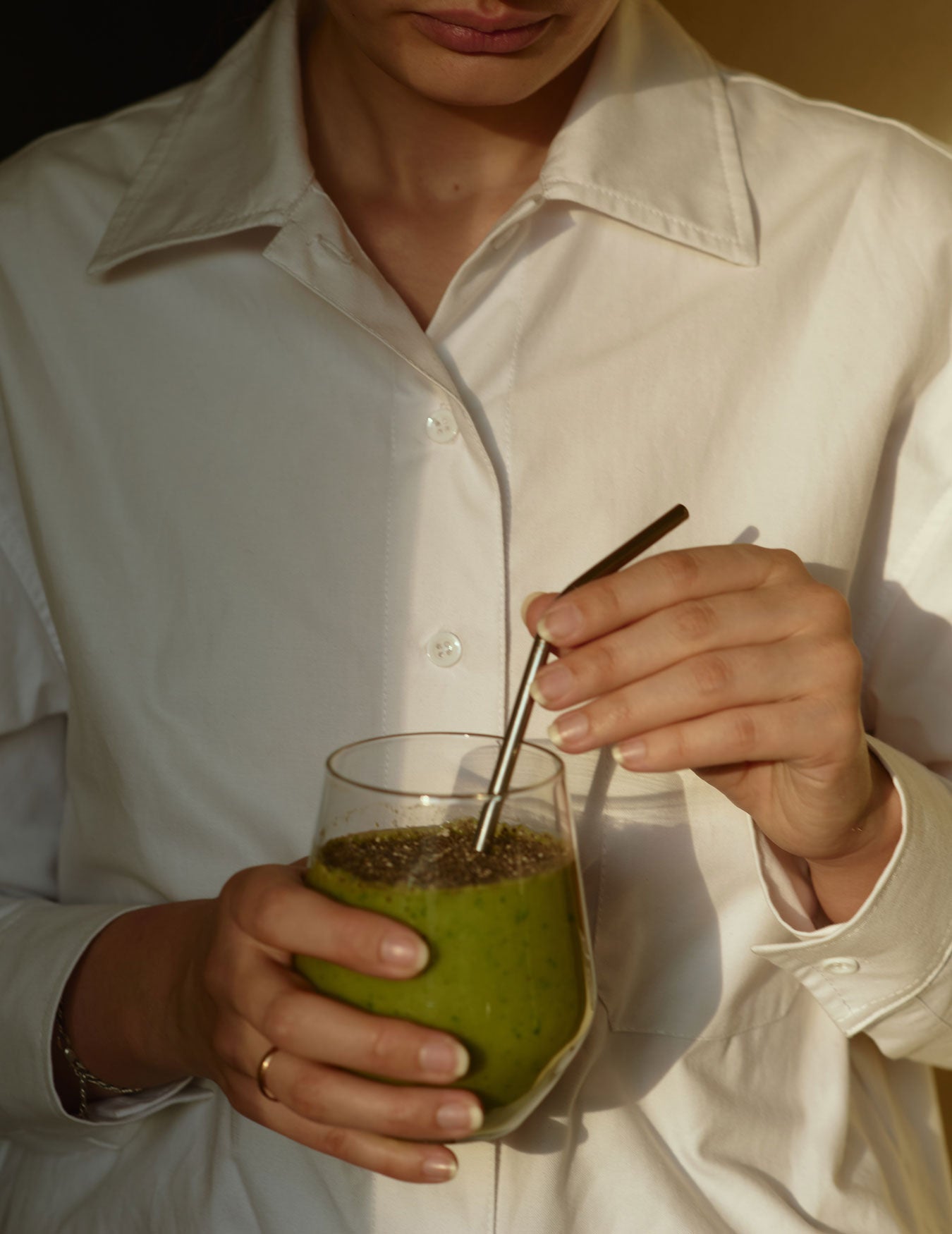 Woman in a white button down holding a glass with a green smoothie and silver straw in her hand
