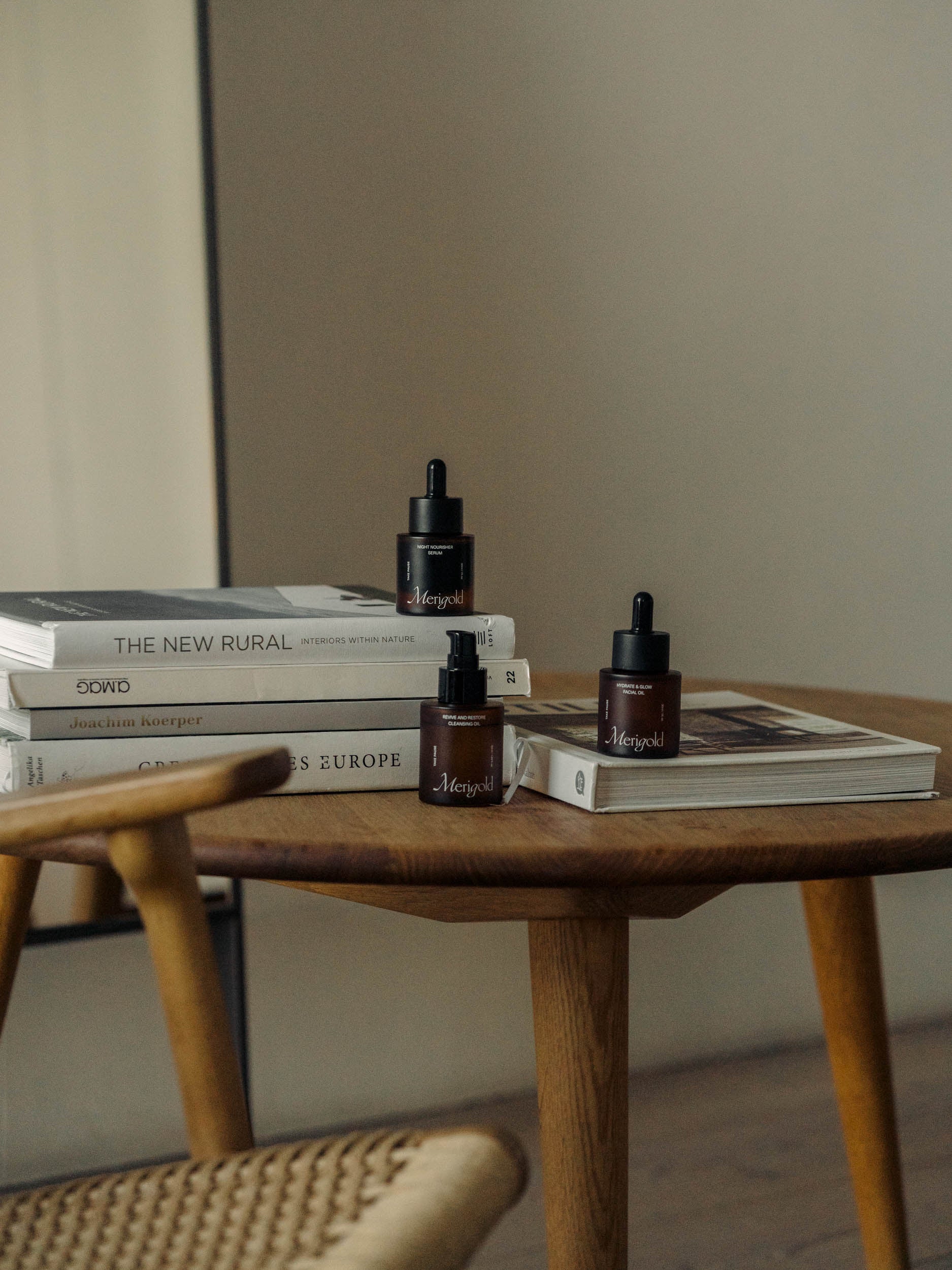 Merigold Oil Trio, including Hydrate and Glow Facial Oil, Revive and Restore Cleansing Oil, and Night Nourisher Serum, styled on a wooden table with books in a cozy, minimalist home setting