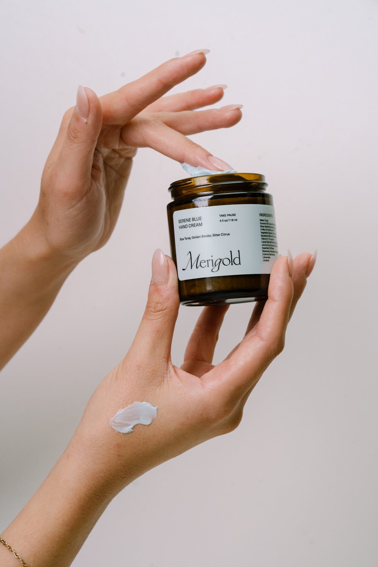 Person applying the Serene Blue hand cream from a jar labeled 'Merigold' to their hand against a neutral background