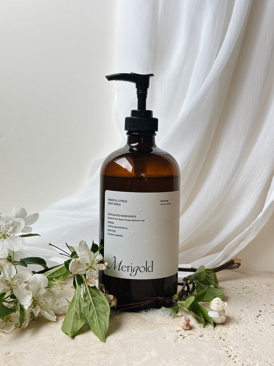 Merigold Mindful Citrus Body Wash styled with delicate flowers on a cream fabric backdrop, uplifting bergamot body cleanser.
