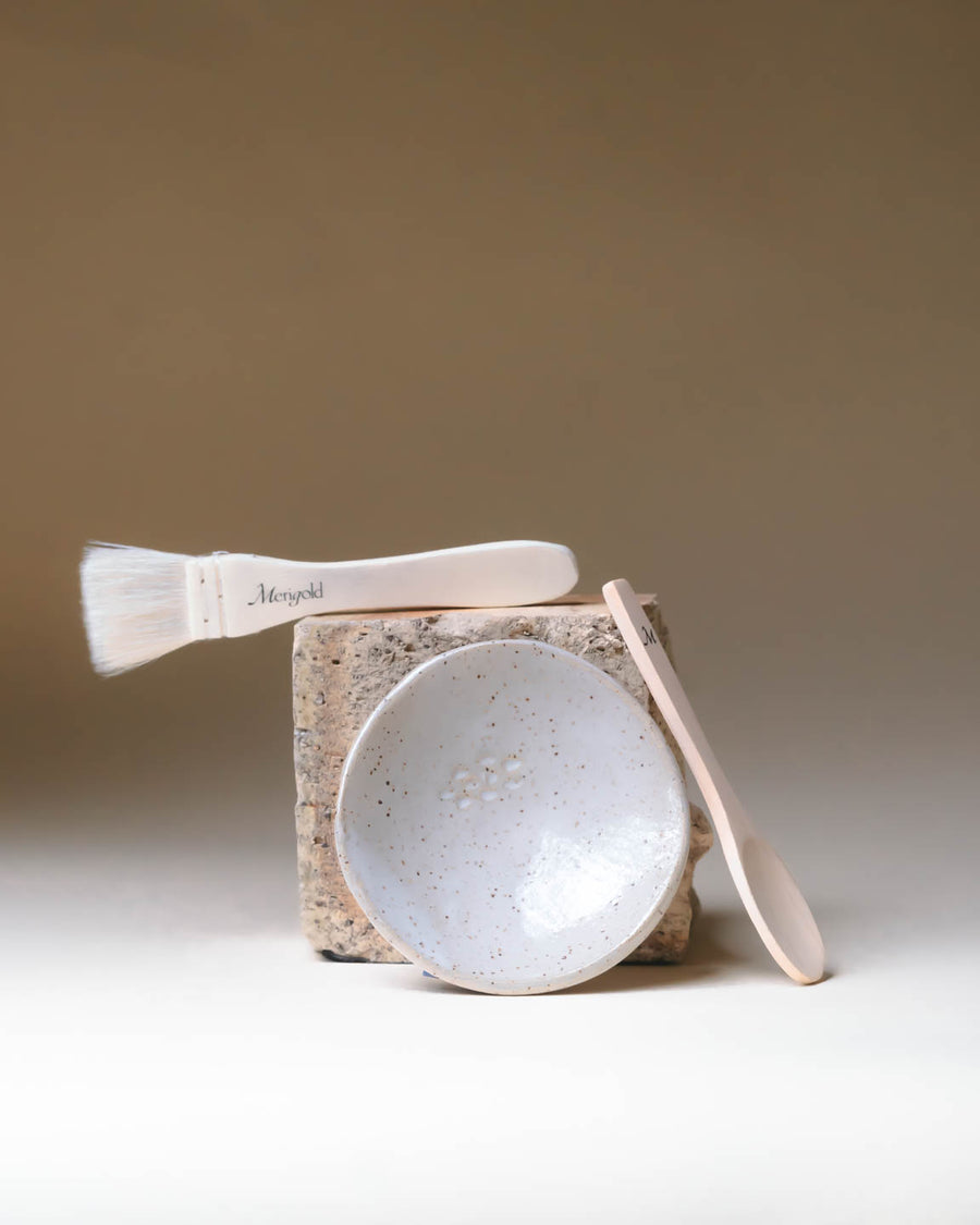 Merigold masking ritual tools—ceramic bowl, wooden spoon, and mask brush—styled on natural stone for clean skincare rituals.