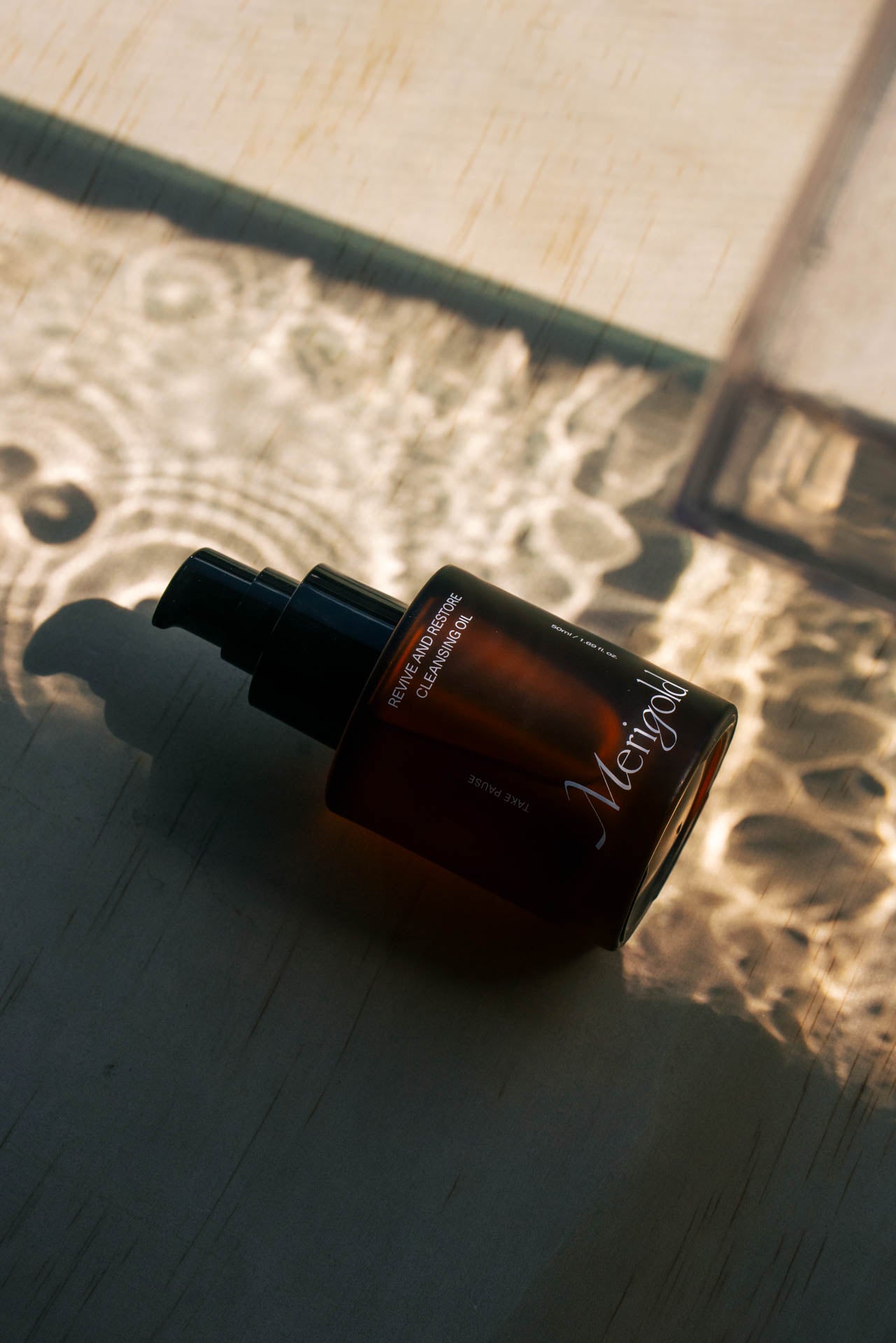 Close-up of Merigold Revive and Restore Cleansing Oil bottle lying on a sunlit surface with artistic light reflections