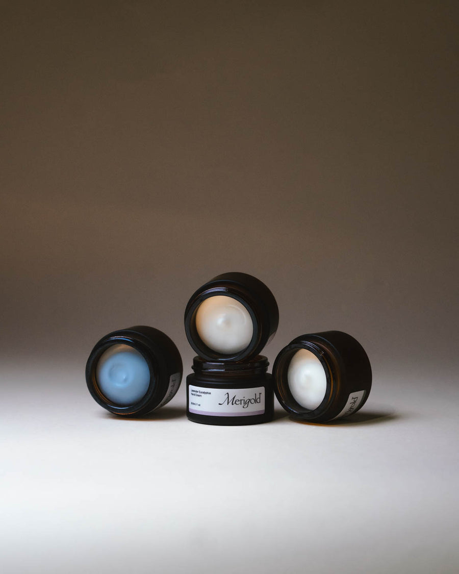 Merigold hand creams displayed open, featuring Serene Blue with Blue Tansy’s soft blue hue, Mindful Citrus hand cream, Woodland Zen hand cream, and Lavender Eucalyptus hand cream. Showcasing rich creamy texture in amber jars.