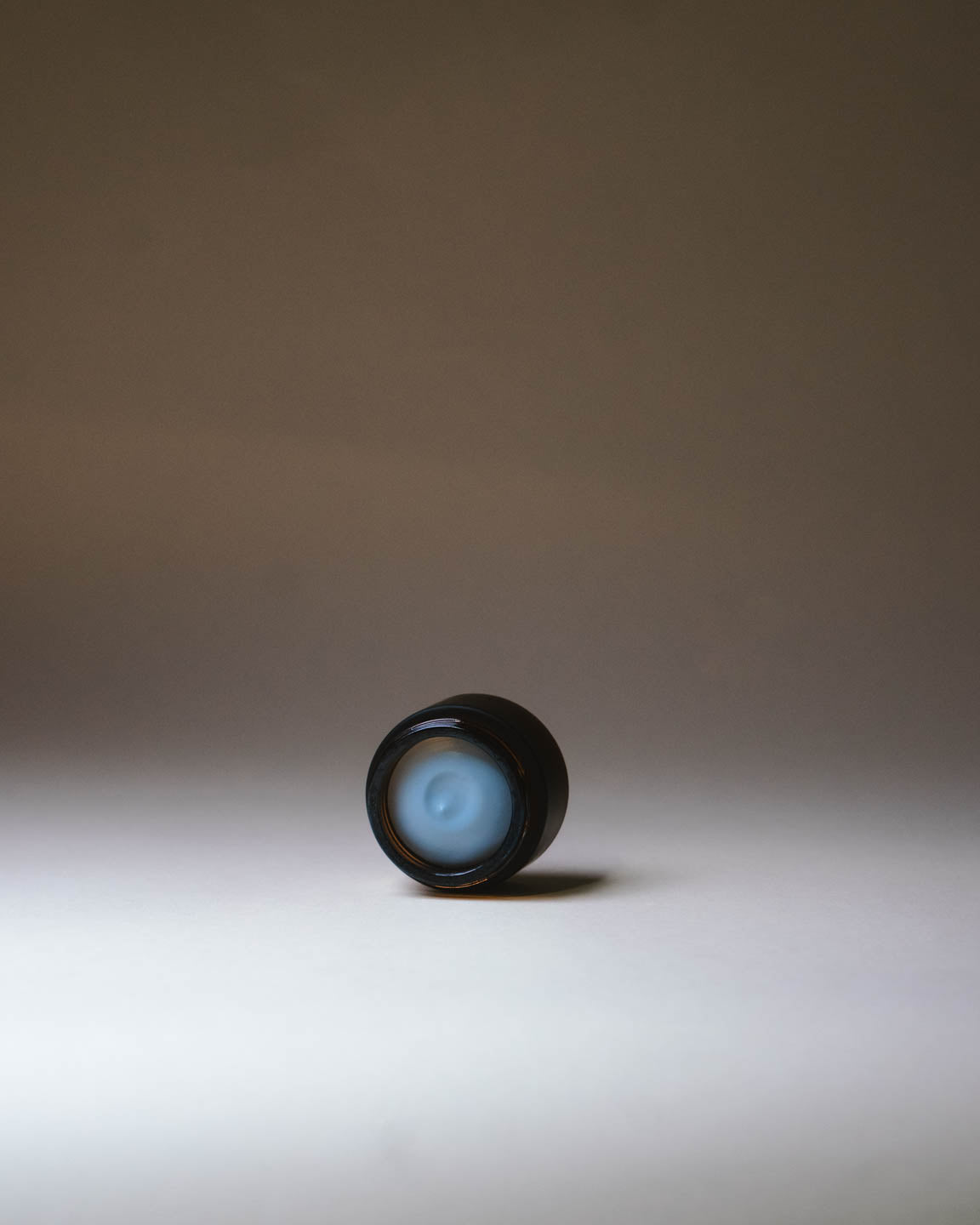 Amber glass jar tipped on it's side with Merigold Serene blue hand cream showing texture on a gradient background.