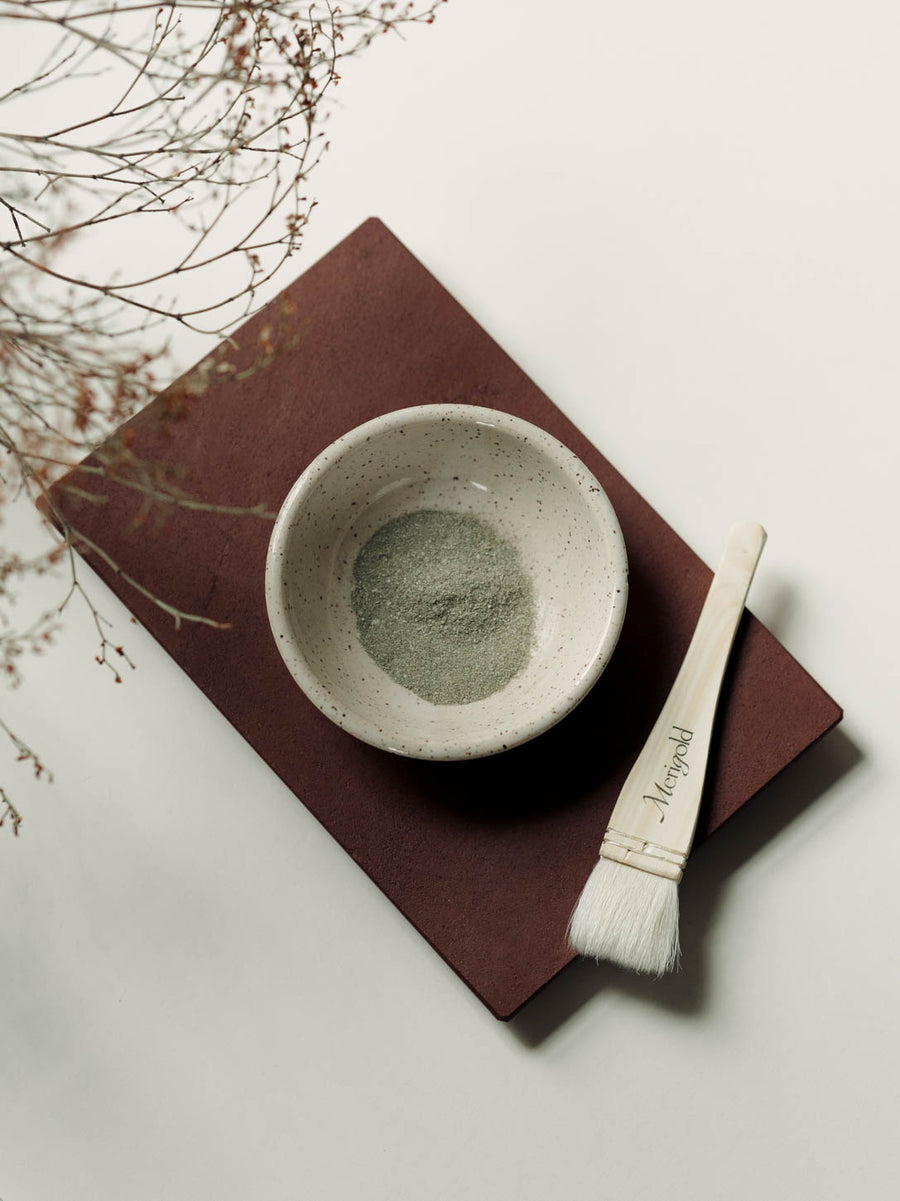 Merigold powdered face mask in handmade ceramic bowl, on red display stone with brush on side. 